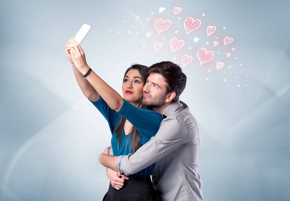 A young couple in love and drawn red hearts taking selfie with a mobile phone in the handsome guys hand in front of an empty clear grey wall background concept.jpeg