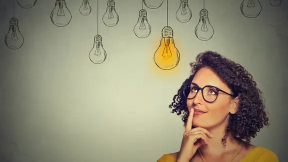 Thinking woman in glasses looking up with light idea bulb above head isolated on gray wall background.jpeg