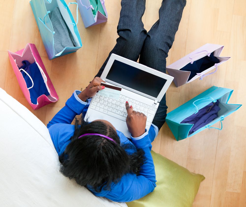 Pretty afro-american teenager using a laptop sitting between shopping bags on the floor in the living room.jpeg