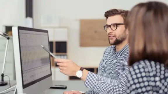 Proficient young male employee with eyeglasses and checkered shirt%2c explaining a business analysis displayed on the monitor of a desktop PC to his female colleague%2c in the interior of a modern office-1.jpeg