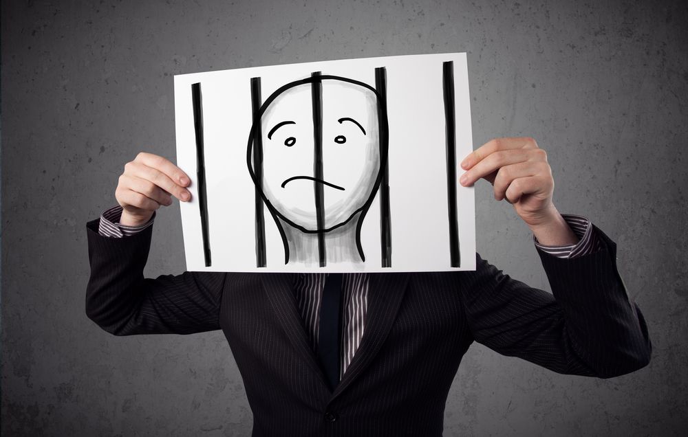 Businessman holding a paper with a prisoner in jail behind the bars on it in front of his head.jpeg