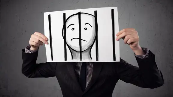 Businessman holding a paper with a prisoner in jail behind the bars on it in front of his head.jpeg