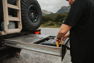 Man pulling an M2 Overland custom tray compartment out of the back of a truck with mountains and wilderness in background 