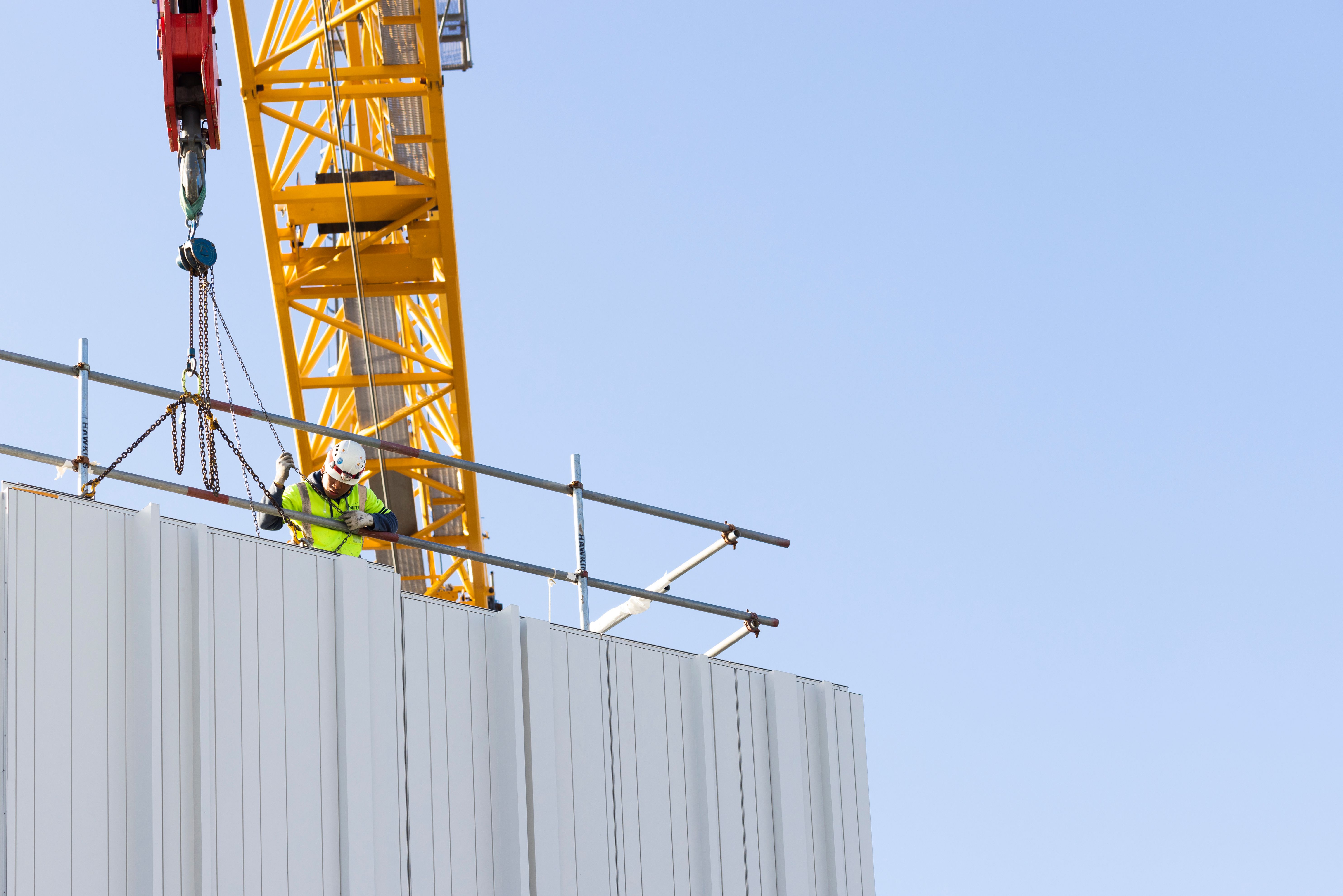 A construction worker securing scaffolding on the corner of a building with a crane overhead 