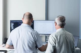 Two men standing in front of a computer, discussing a CAD model on the screen 