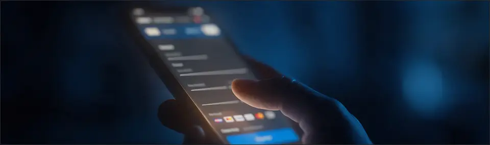 A hand holding a smartphone showing a checkout form with payment method icons, in a dark room.