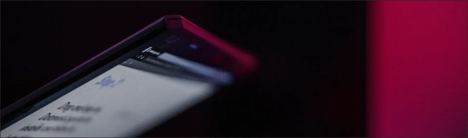 Close-up of the edge of a smartphone lying on a surface, photographed from a low angle. The device's screen displays bright content with illegible text and an icon. The photo is captured in a dark, atmospheric tone with a dominant pink-red backlight in the background.