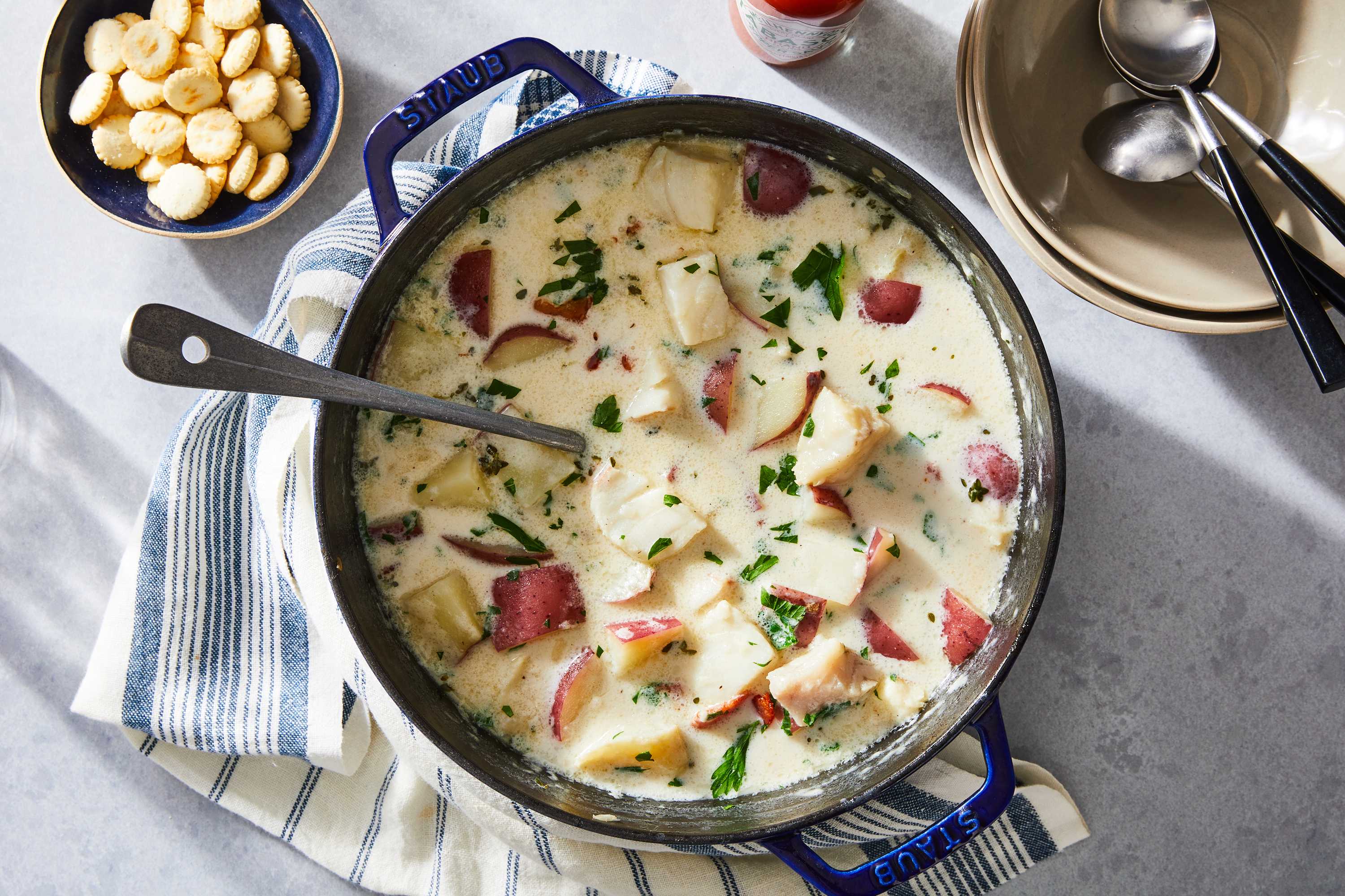 a pot of potato and fish soup on a table with spoons, bowls, and oyster crackers