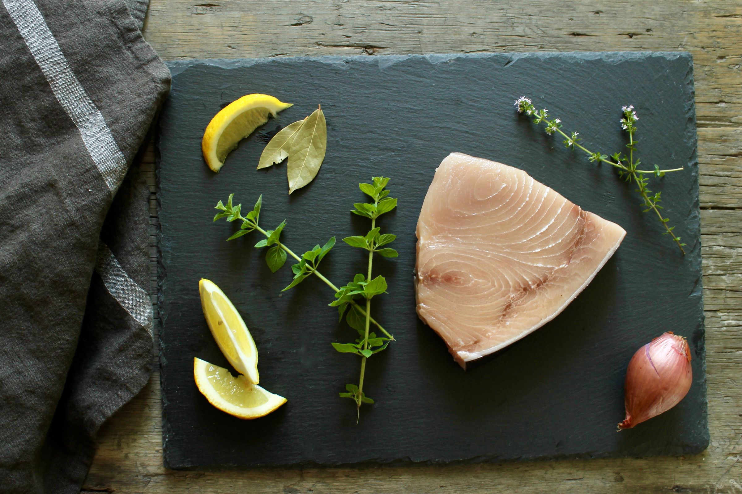 a slate cutting board topped with a piece of swordfish, lemon slices, onions and herbs .