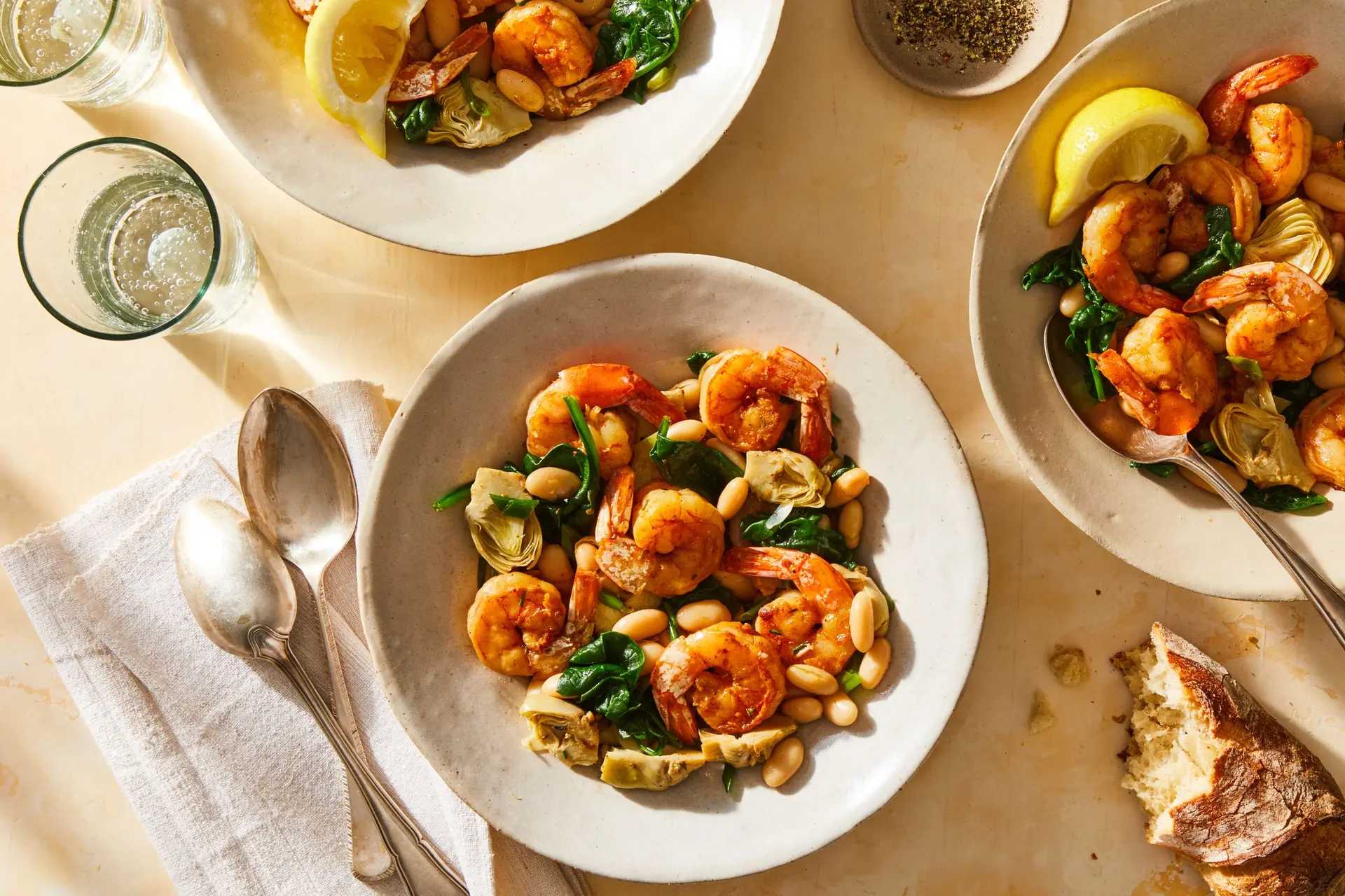 three bowls of shrimp on a table with spoons, bread, and glasses