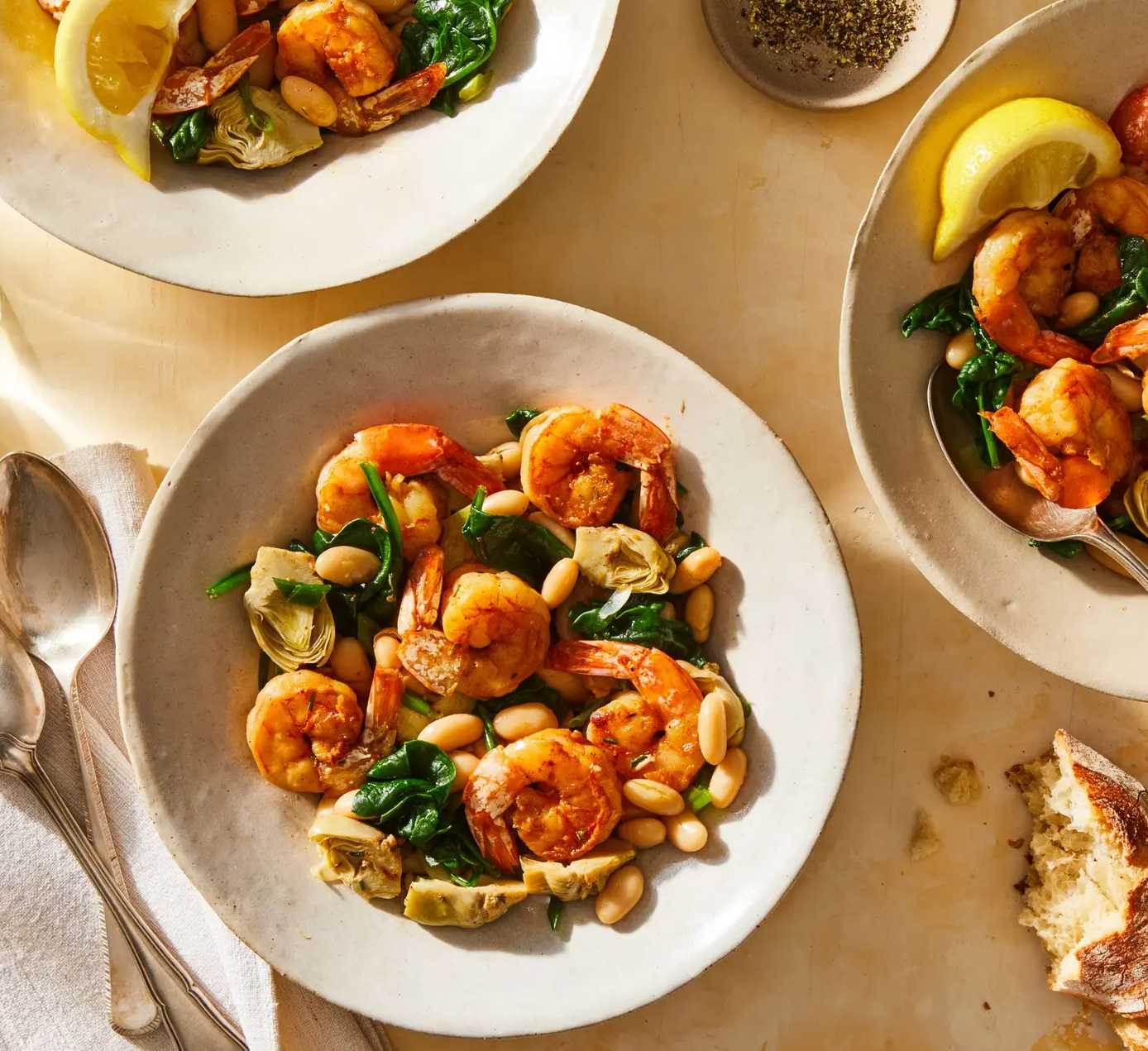 three bowls of shrimp on a table with spoons, bread, and glasses