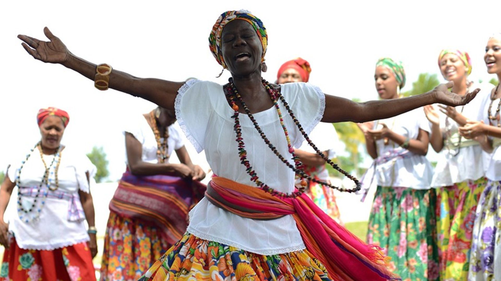 Ganhadeira Raquel das Virgens aparece em primeiro plano, cantando de braços abertos, com outras ganhadeiras em segundo plano