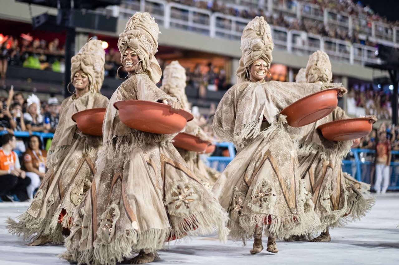 Mulheres com fantasia em tons terrosos, seguram bacias cor de barro em referência ao trabalho das lavadeiras