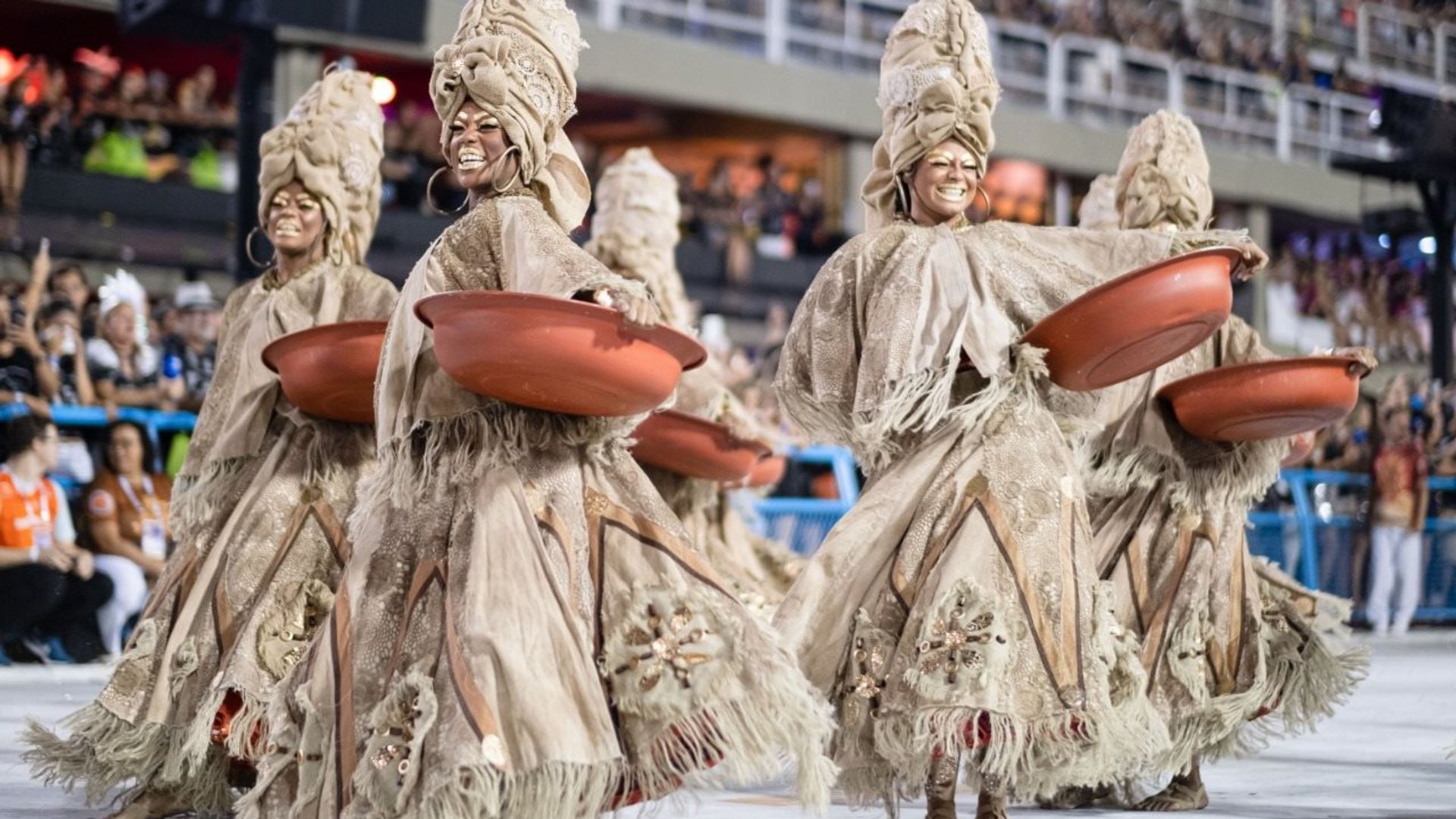 Mulheres com fantasia em tons terrosos, seguram bacias cor de barro em referência ao trabalho das lavadeiras