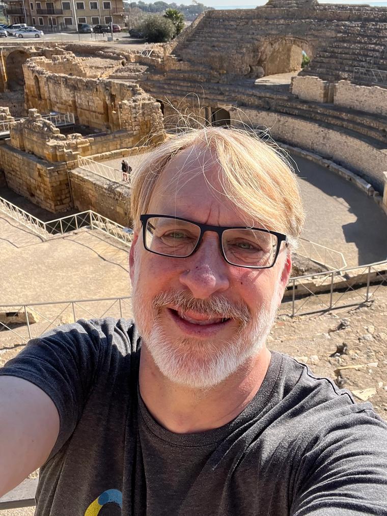 Rusty Cole taking a selfie in front of ancient Roman amphitheater ruins with the sea in the distance.