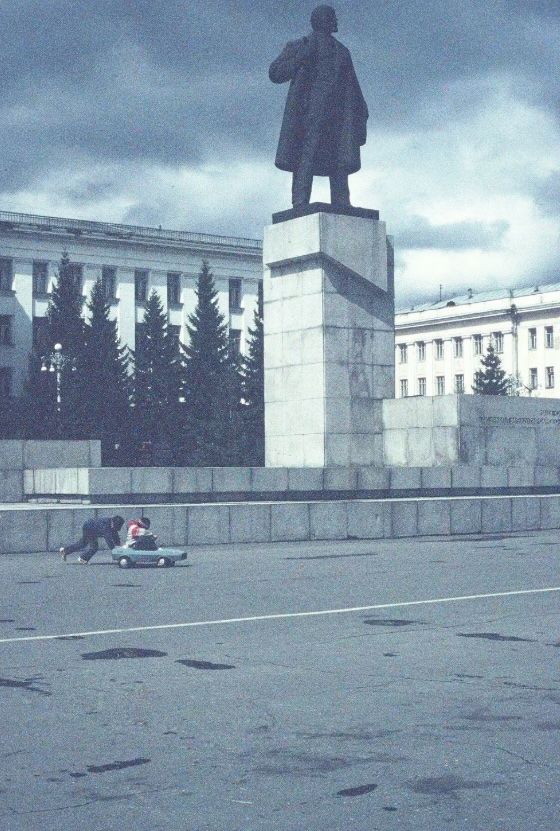 A dark statue of Lenin overlooks a child pushing another child in a toy car in a plaza under a cloudy sky.