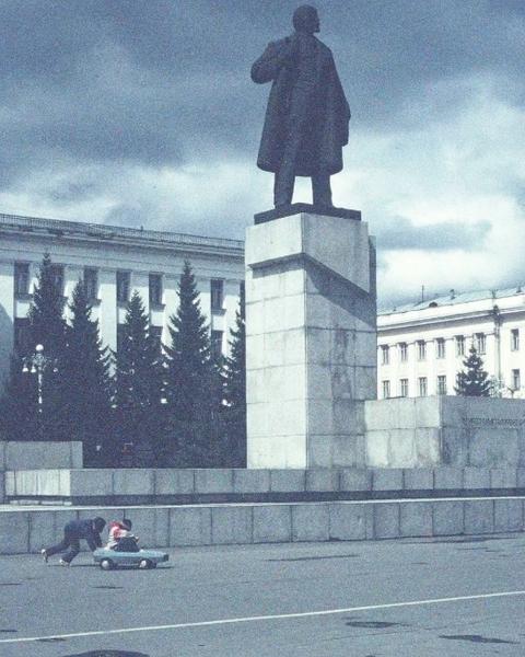A dark statue of Lenin overlooks a child pushing another child in a toy car in a plaza under a cloudy sky.