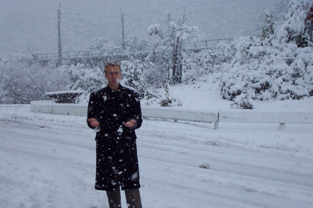 Rusty Cole standing in heavy snowfall in a long black coat.