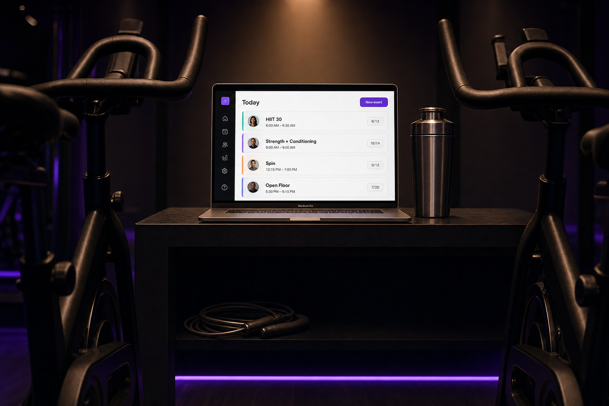 Cinematic shot between two matte-black spin bikes toward a MacBook on a charcoal counter displaying the Vibefam schedule (HIIT 30, Strength + Conditioning, Spin, Open Floor) with a chrome shaker bottle beside it; deep purple LED lighting at floor level.