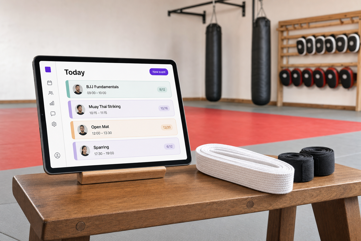 Floor-level view of an iPad on a wooden bench at the edge of a tatami mat displaying the Vibefam schedule (BJJ Fundamentals, Muay Thai Striking, Open Mat, Sparring), beside a folded white belt and coiled black hand-wraps, with heavy bags hanging in the soft-focus background.
