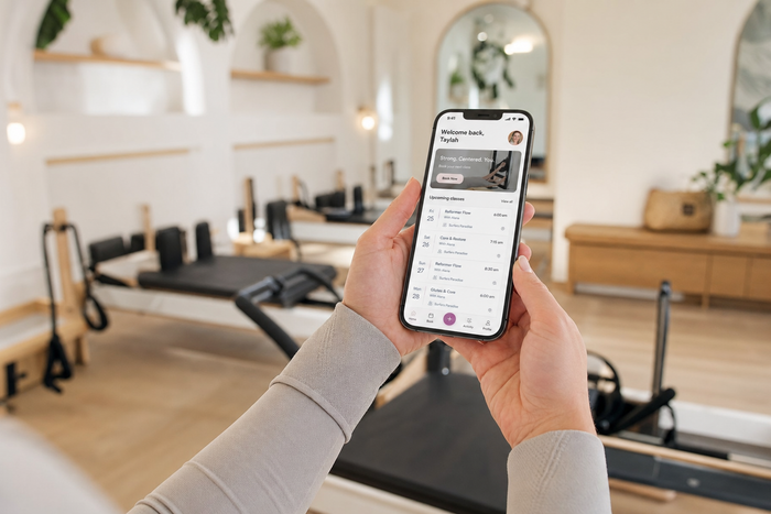 Hands holding an iPhone displaying a boutique studio's custom-branded member app with a bookings list, soft-focus modern pilates reformer studio in the background.