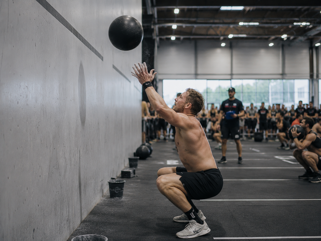 HYROX athlete mid wall-ball rep in a deep squat, ball chalked and mid-throw at a wall target, race arena crowd in the background