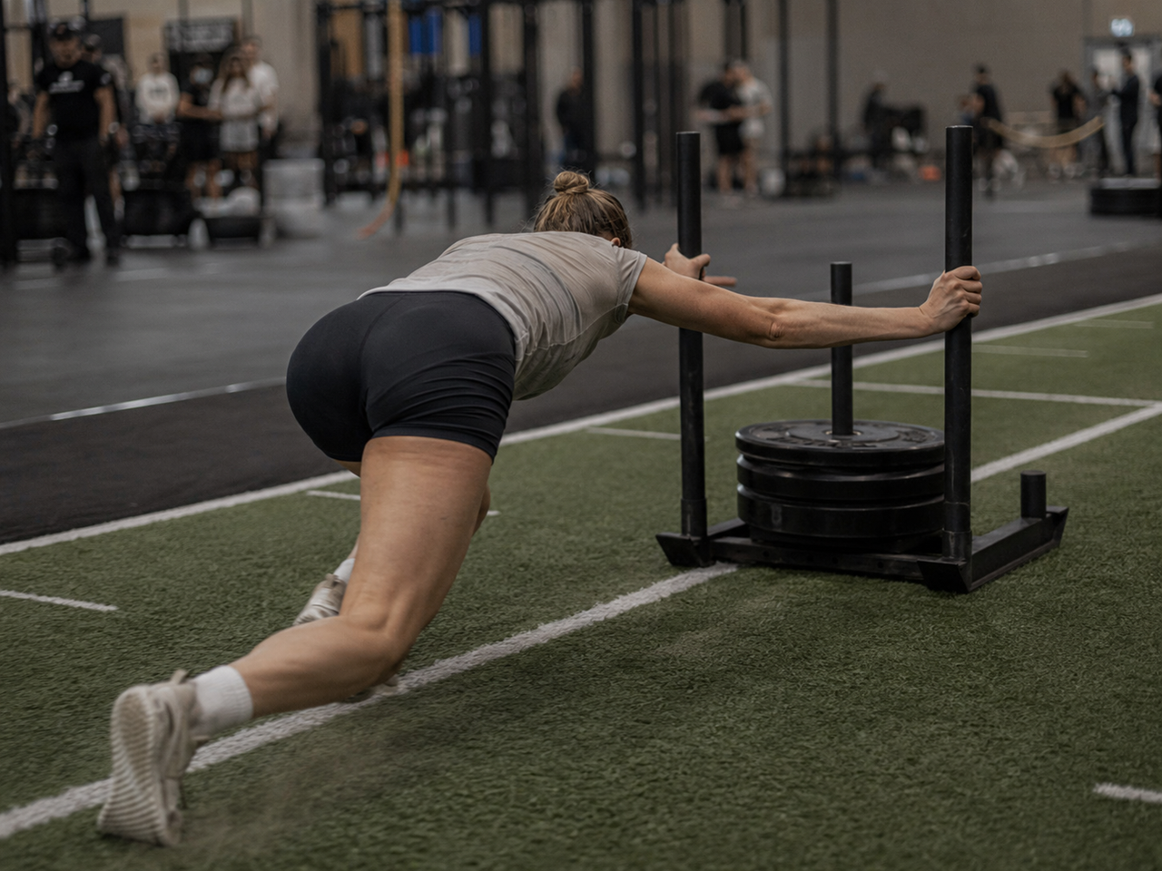 HYROX athlete pushing a loaded sled with a low body position, driving from the legs across turf at a race venue