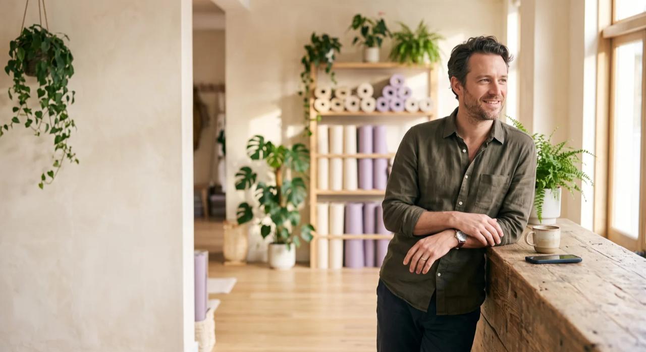 A boutique studio owner leaning on his reception counter, relaxed, his phone face-down beside a coffee