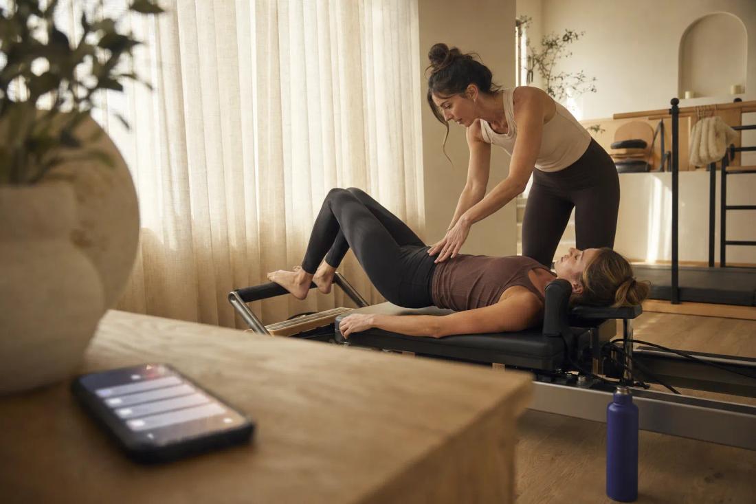 A Pilates studio owner coaches a client on a reformer while her phone, face-up on a side table, handles member messages.
