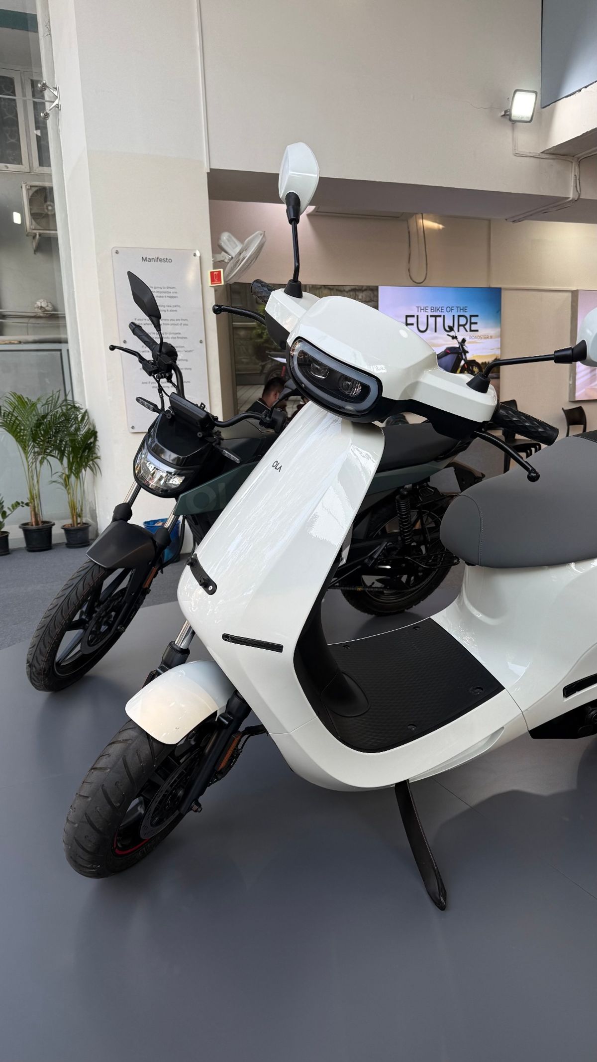 White Ola electric scooter with Roadster motorcycle in background