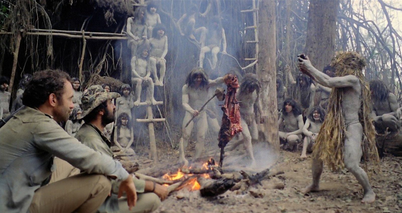 Film still: two men in khaki sit by a fire in a jungle clearing, watching grey-painted villagers, one in a grass costume.
