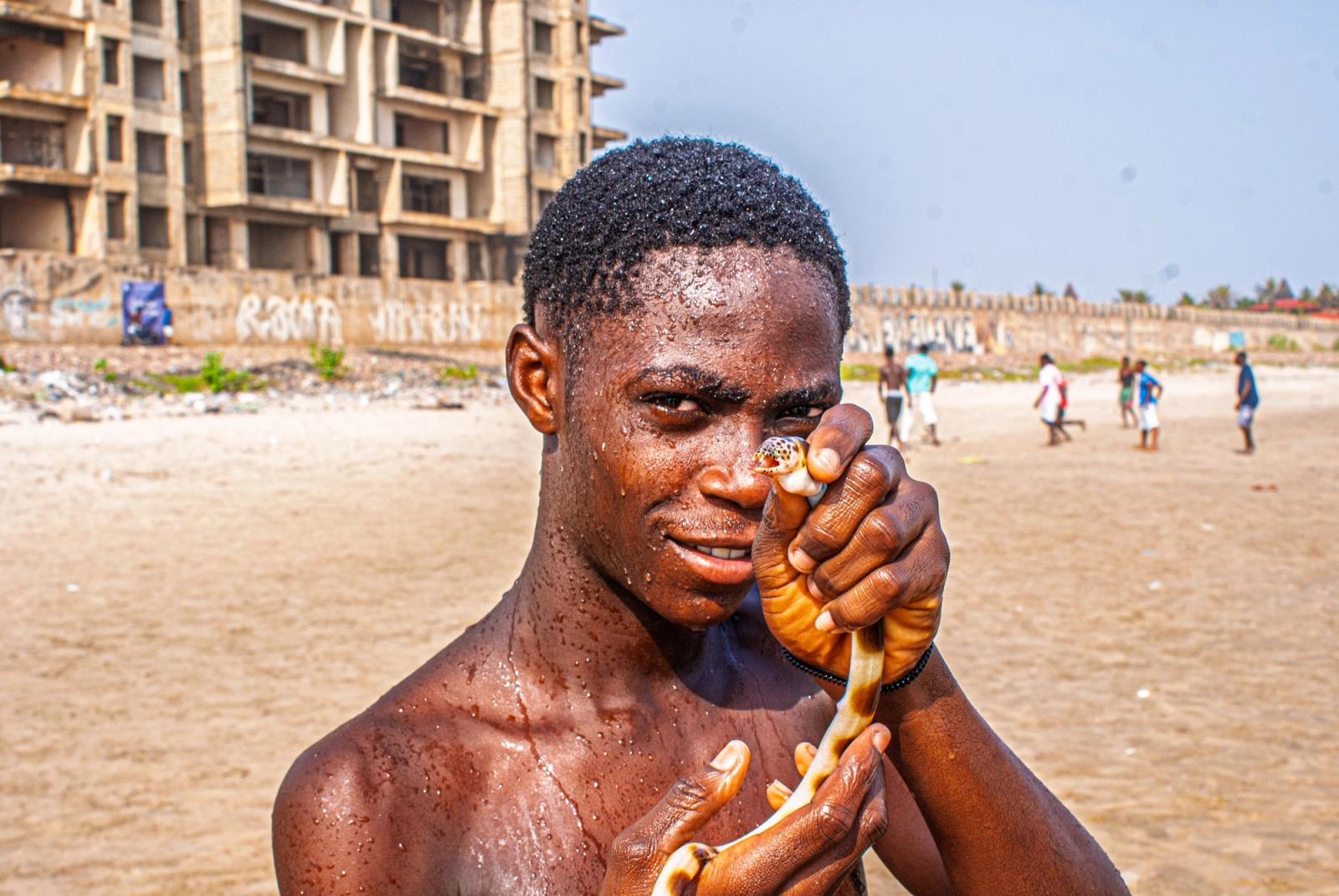 A young man on a beach holds a shell to his face and looks at the camera, with an unfinished concrete building behind him.