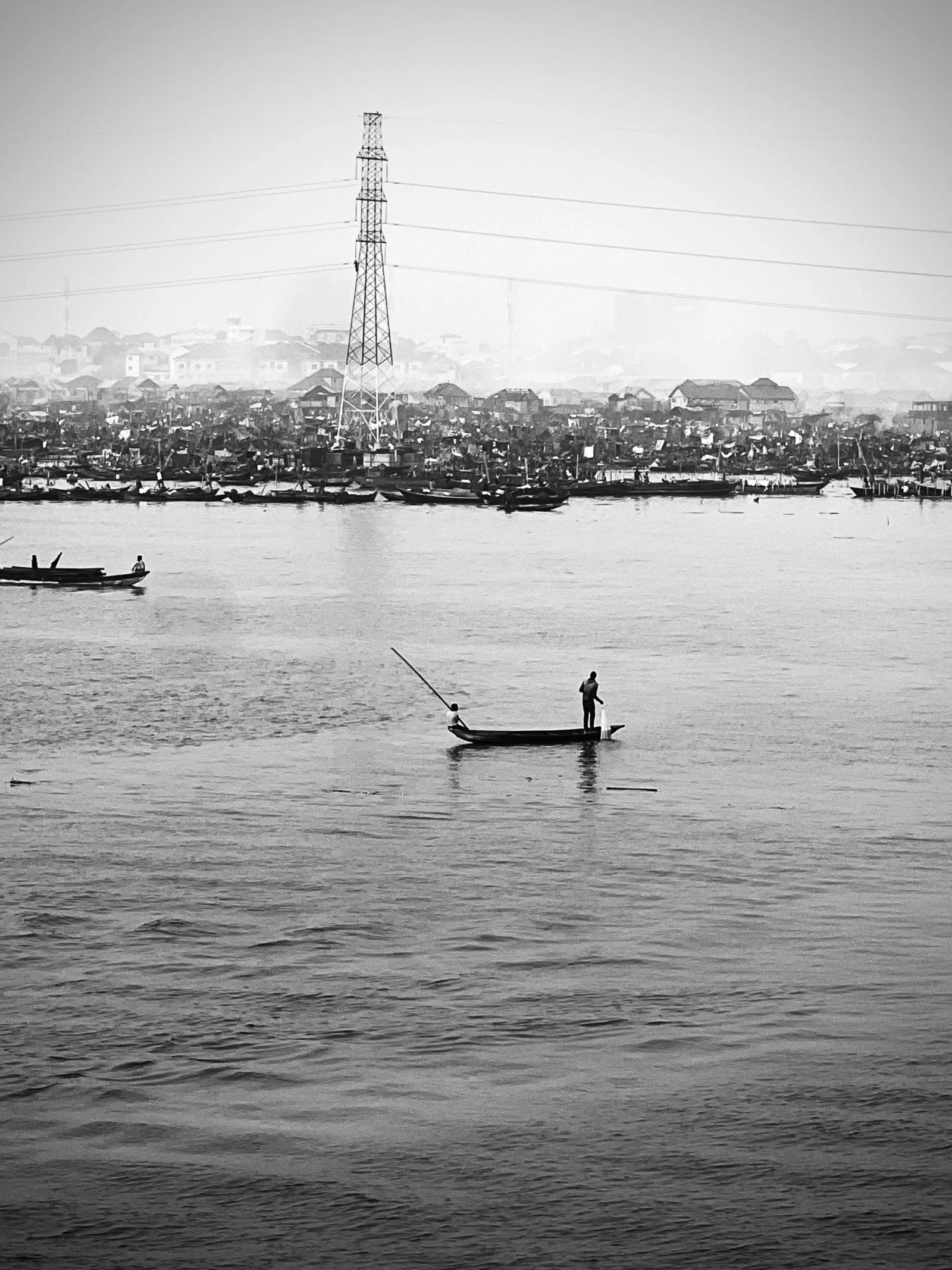 Black-and-white view across a wide lagoon: a man poles a narrow canoe, with a crowded stilted waterfront and an electricity pylon beyond.