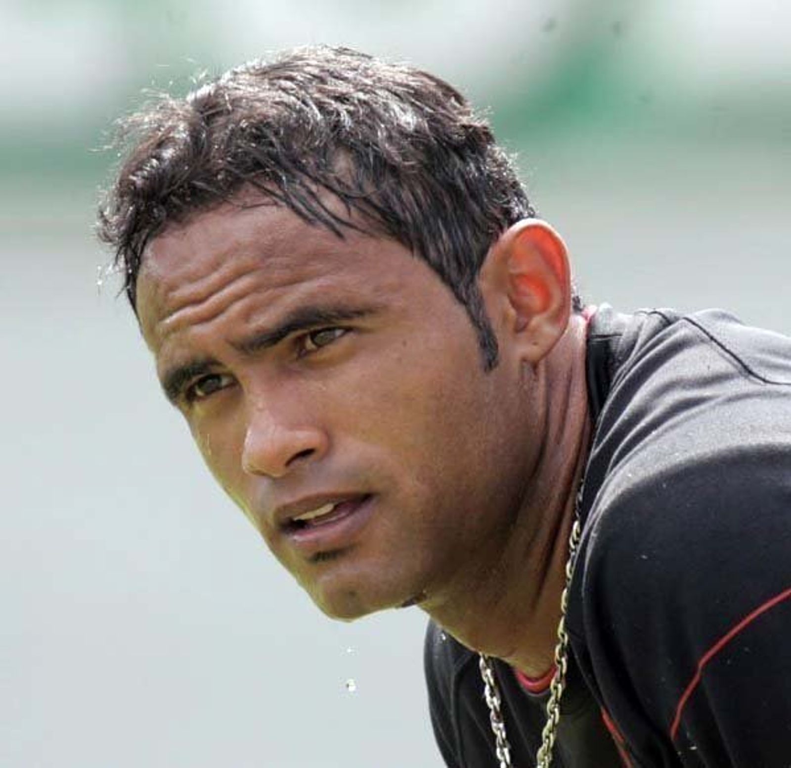Close-up of a man with sweat-damp dark hair and a gold chain, in a dark training top, looking into the distance.