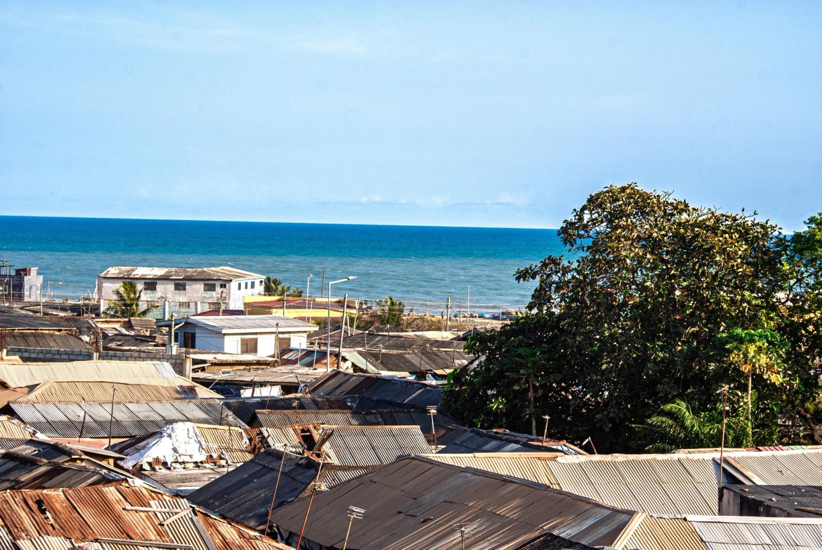 Rusting corrugated roofs of a coastal neighbourhood in the foreground, with the blue sea on the horizon.