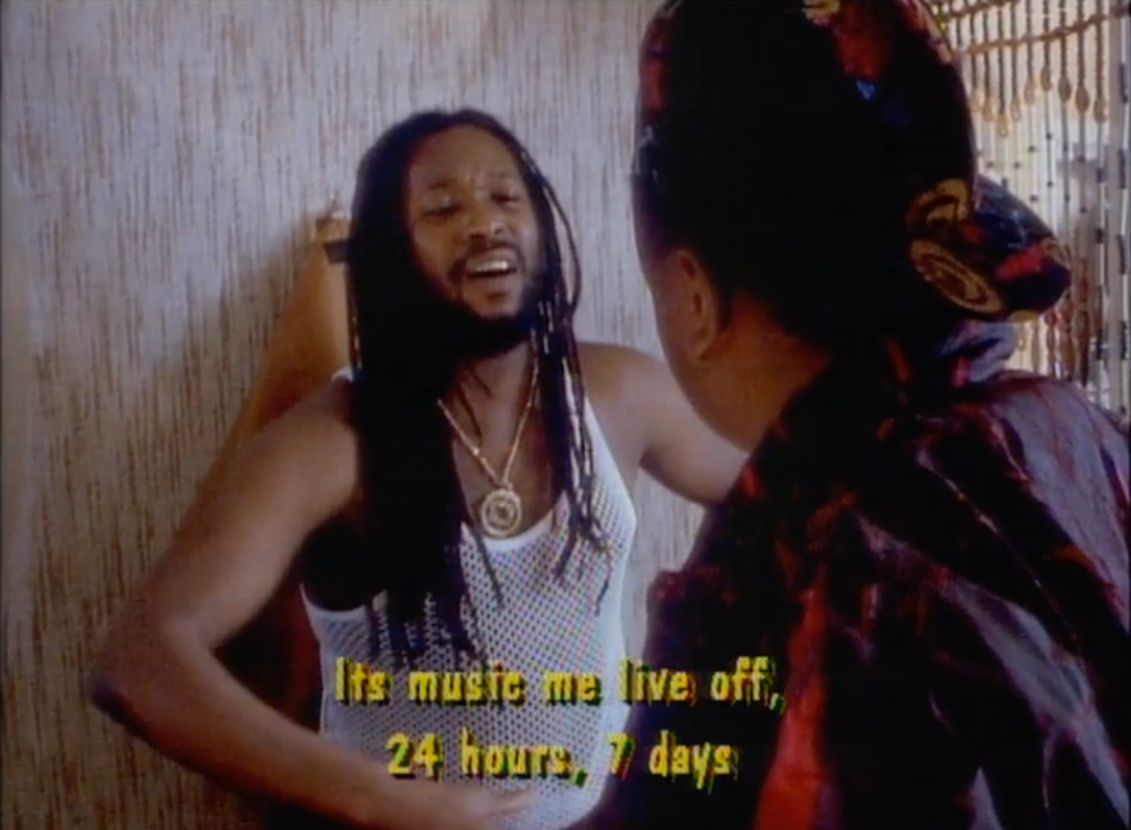 Film still from We The Ragamuffin: a man with long locs in a white mesh vest talks to a woman in a head wrap, with subtitles.