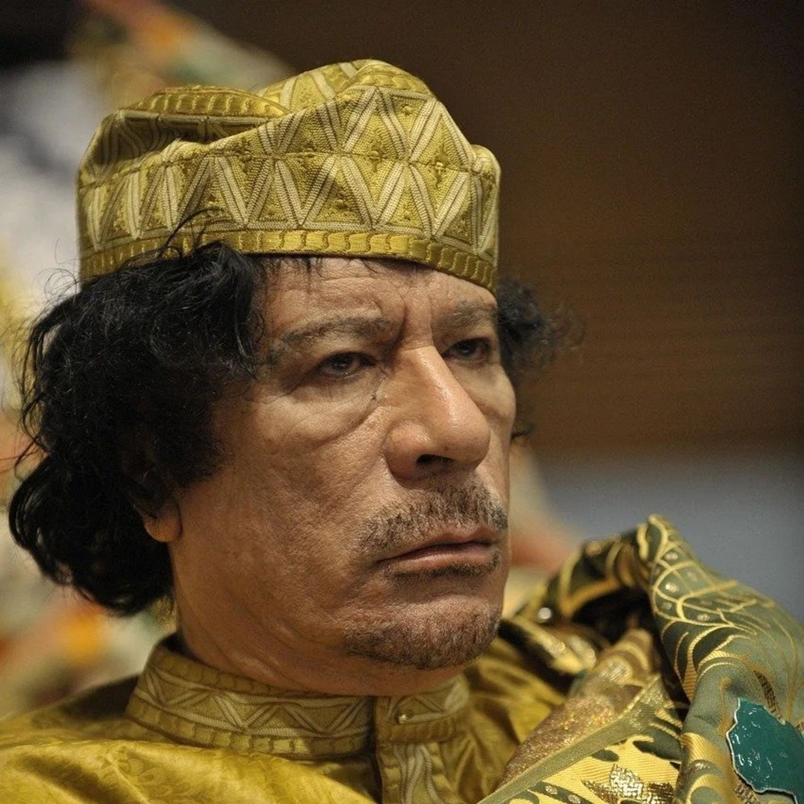 A man with dark curly hair in a gold embroidered cap and a gold patterned robe looks to one side with a stern expression.