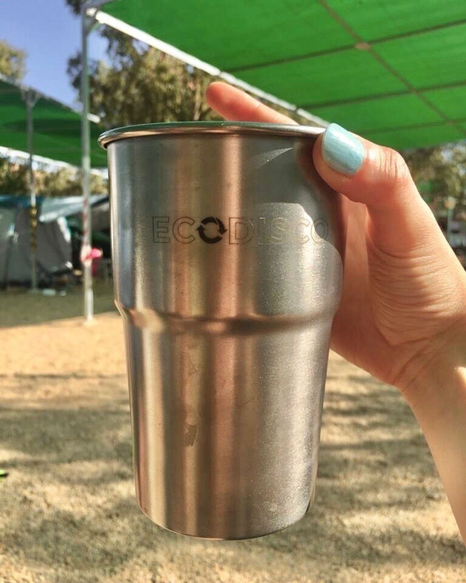 A hand holds a stainless-steel cup engraved with the Ecodisco logo, in a sunny outdoor camp with a green canopy.