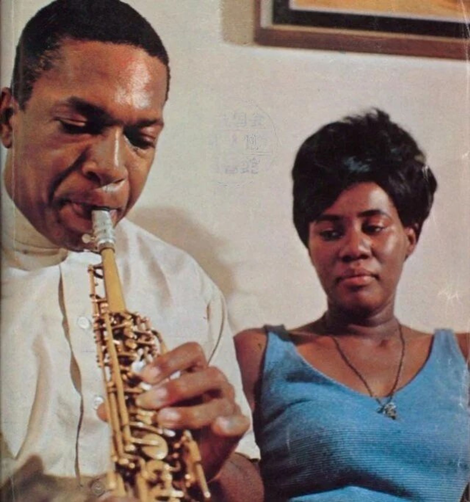 Vintage colour photograph of a man playing a soprano saxophone, eyes lowered, with a woman in a blue top beside him.