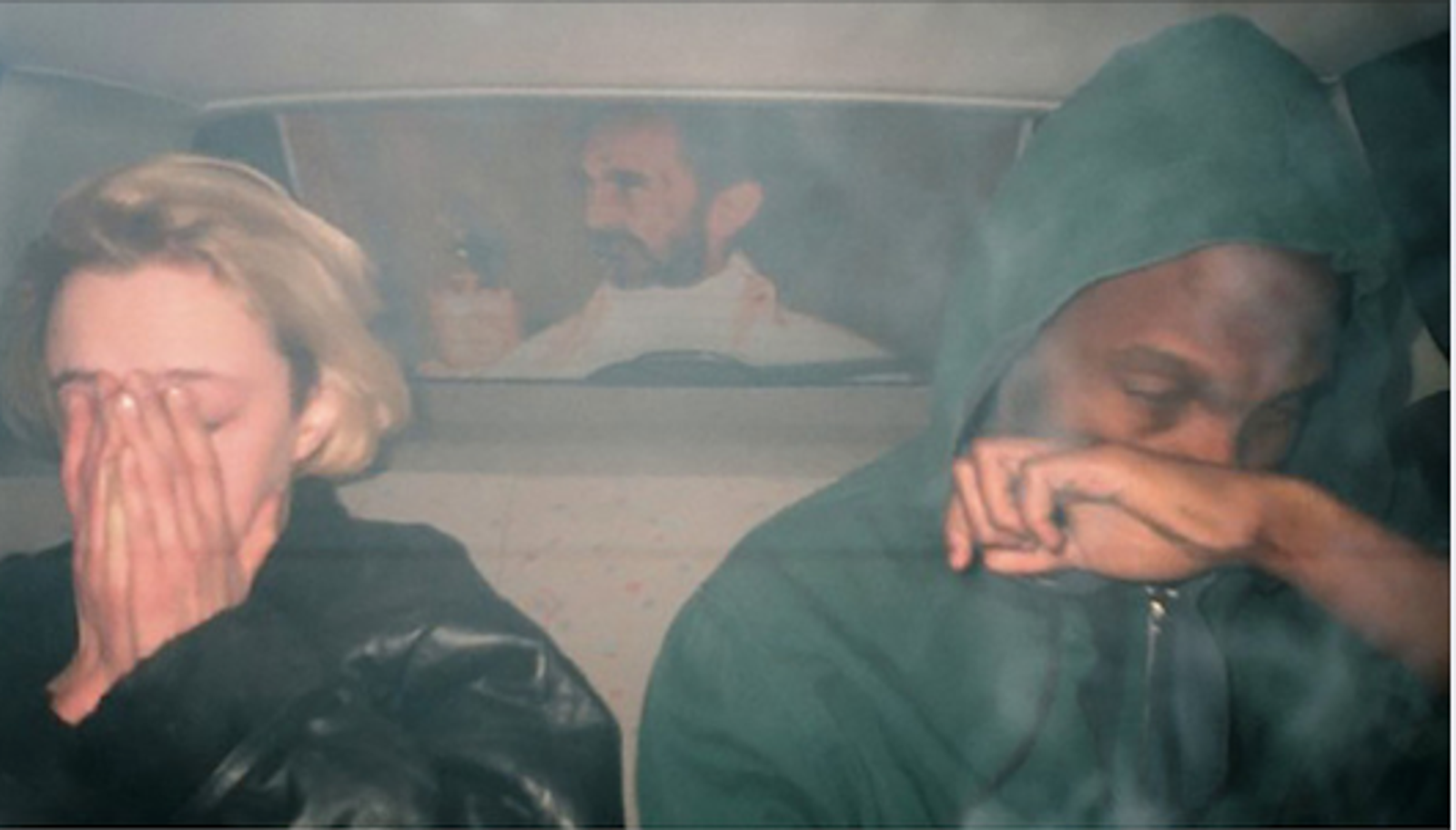 Two people in the back of a car: a blonde woman with her hands over her face and a person in a green hood with a hand over their mouth.