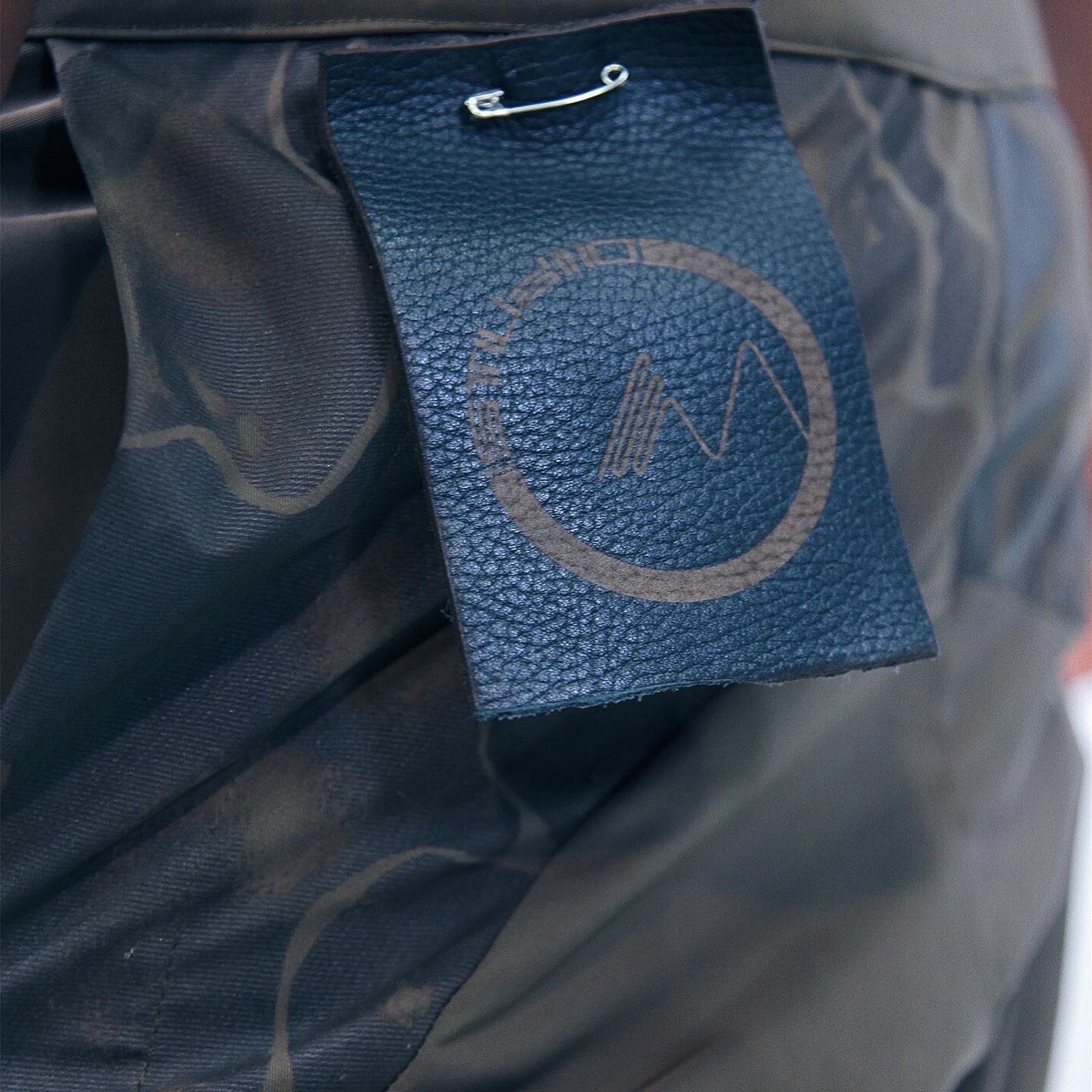 Close-up of a leather label with a circular M logo, pinned to dark olive fabric.