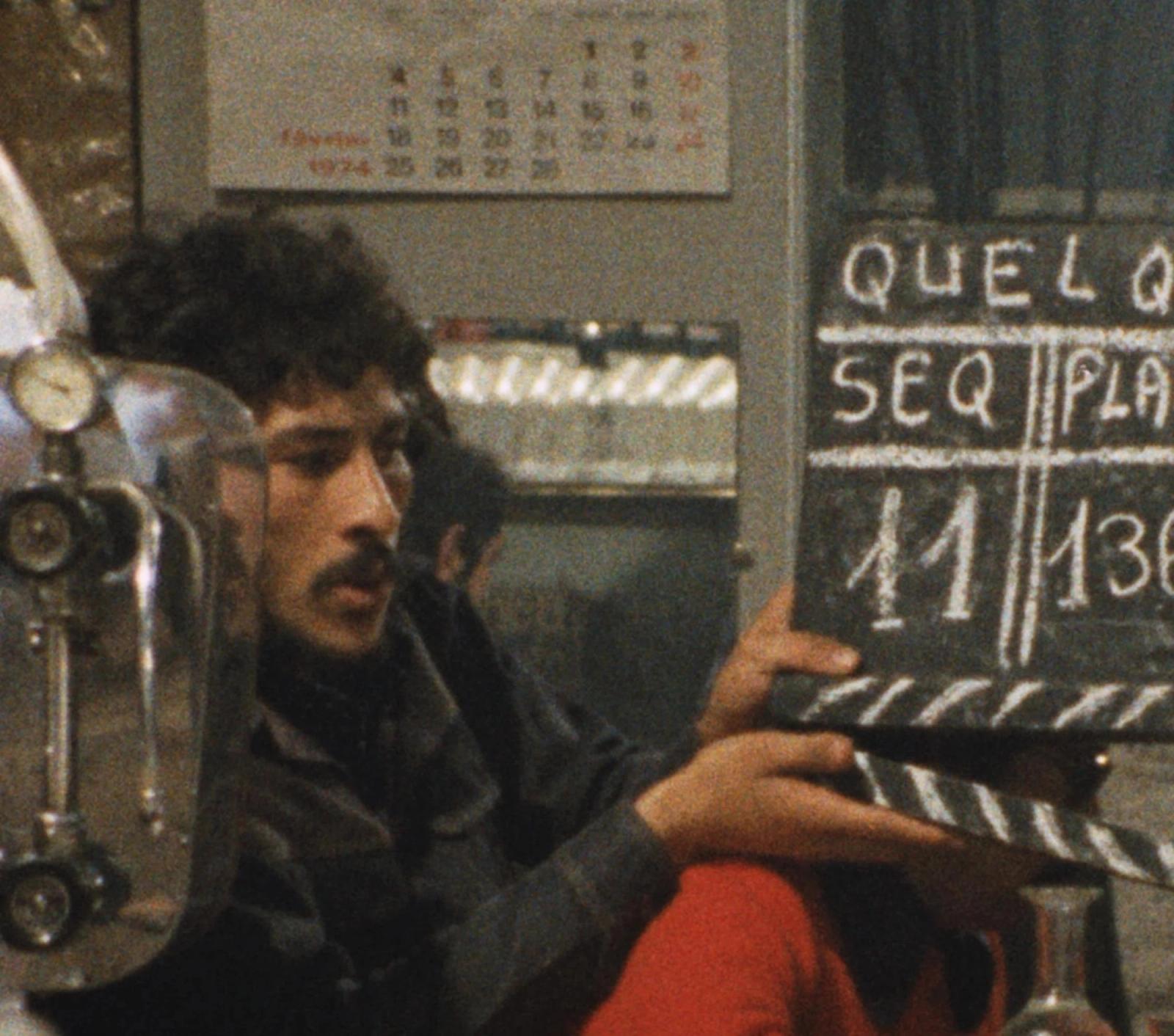 A young man with curly dark hair holds up a clapperboard beside a film camera, with a calendar on the wall behind him.