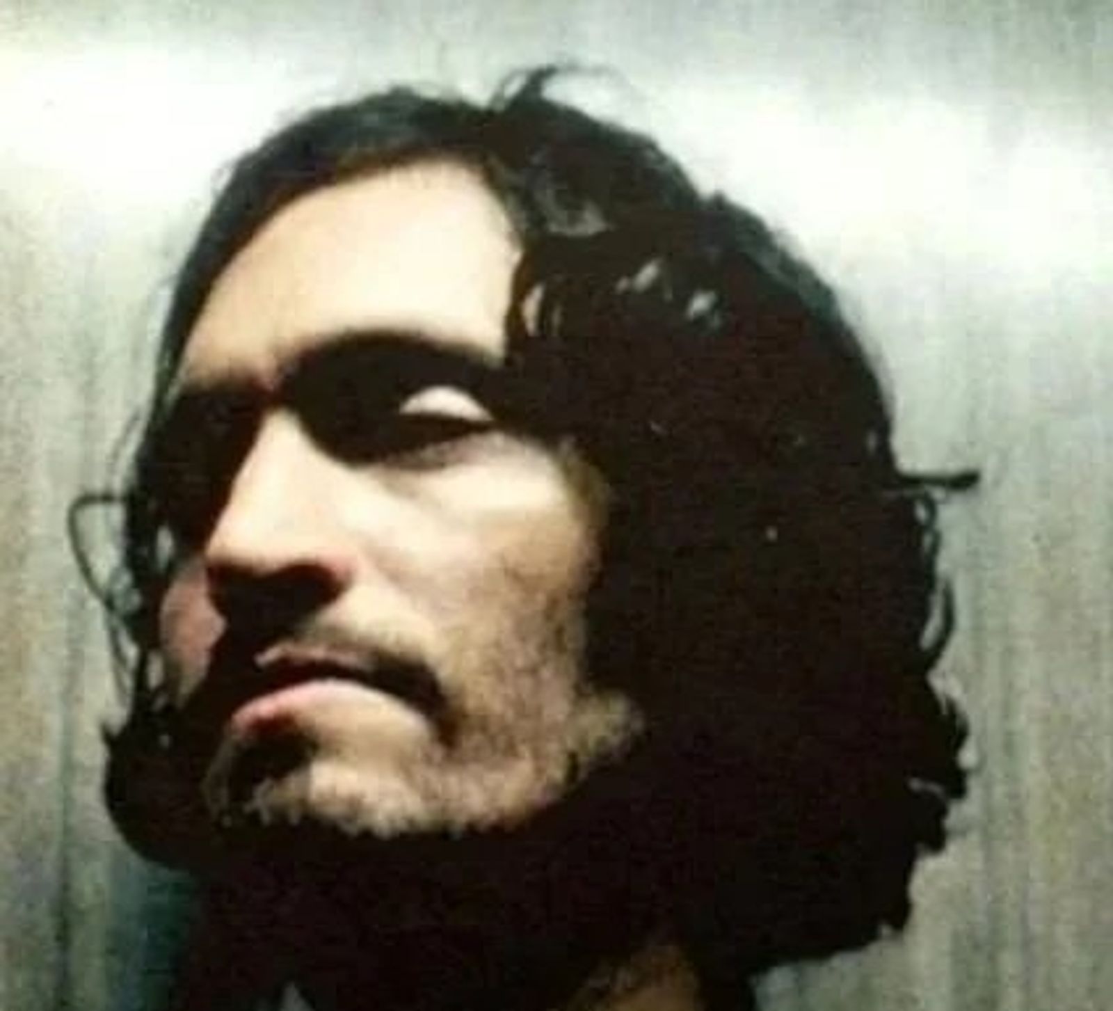 Grainy close-up of a man with long dark hair and a beard, his face half in shadow.