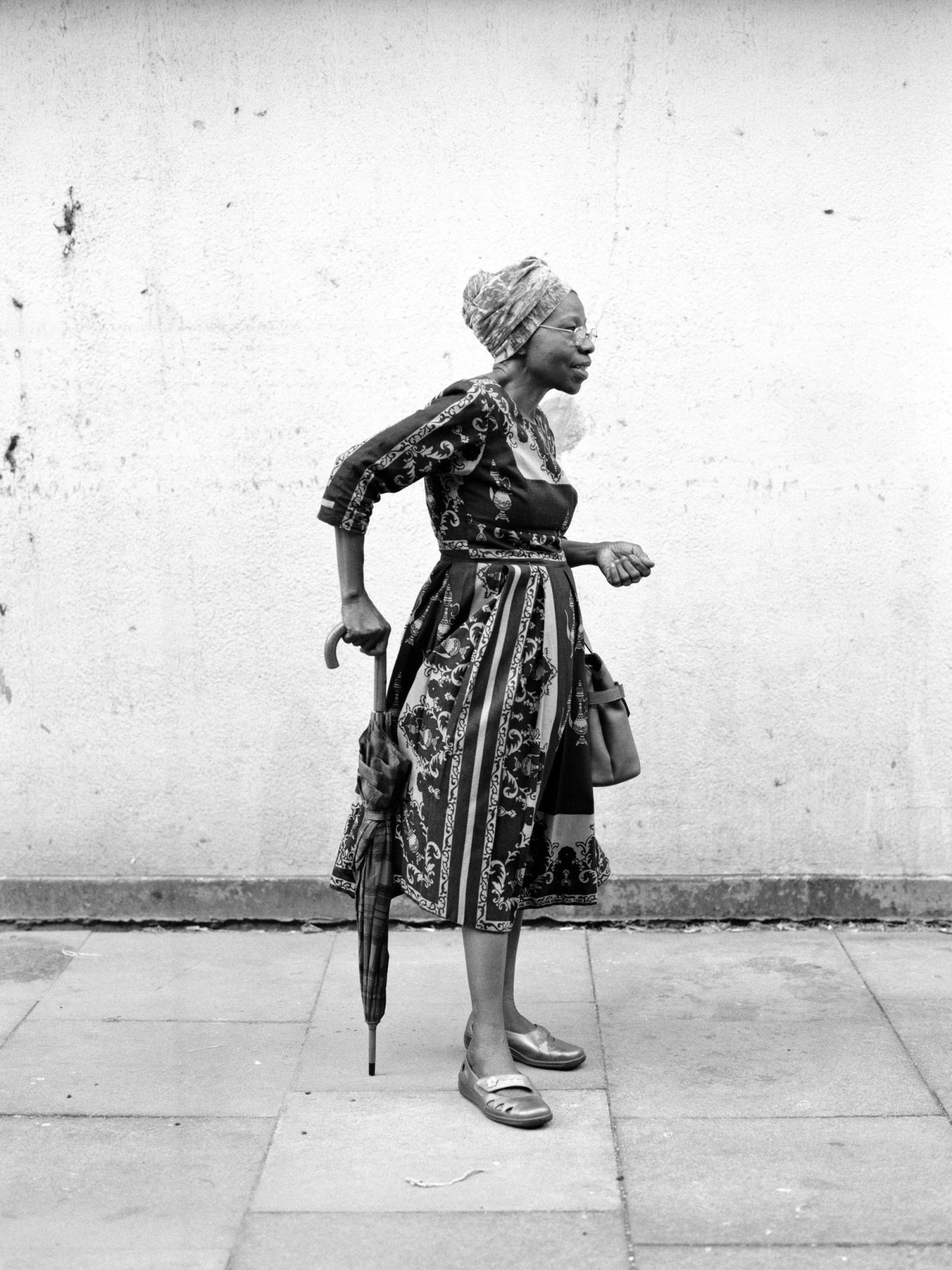 Black-and-white photograph of an older woman in a patterned dress and head wrap, walking in profile and leaning on a closed umbrella.