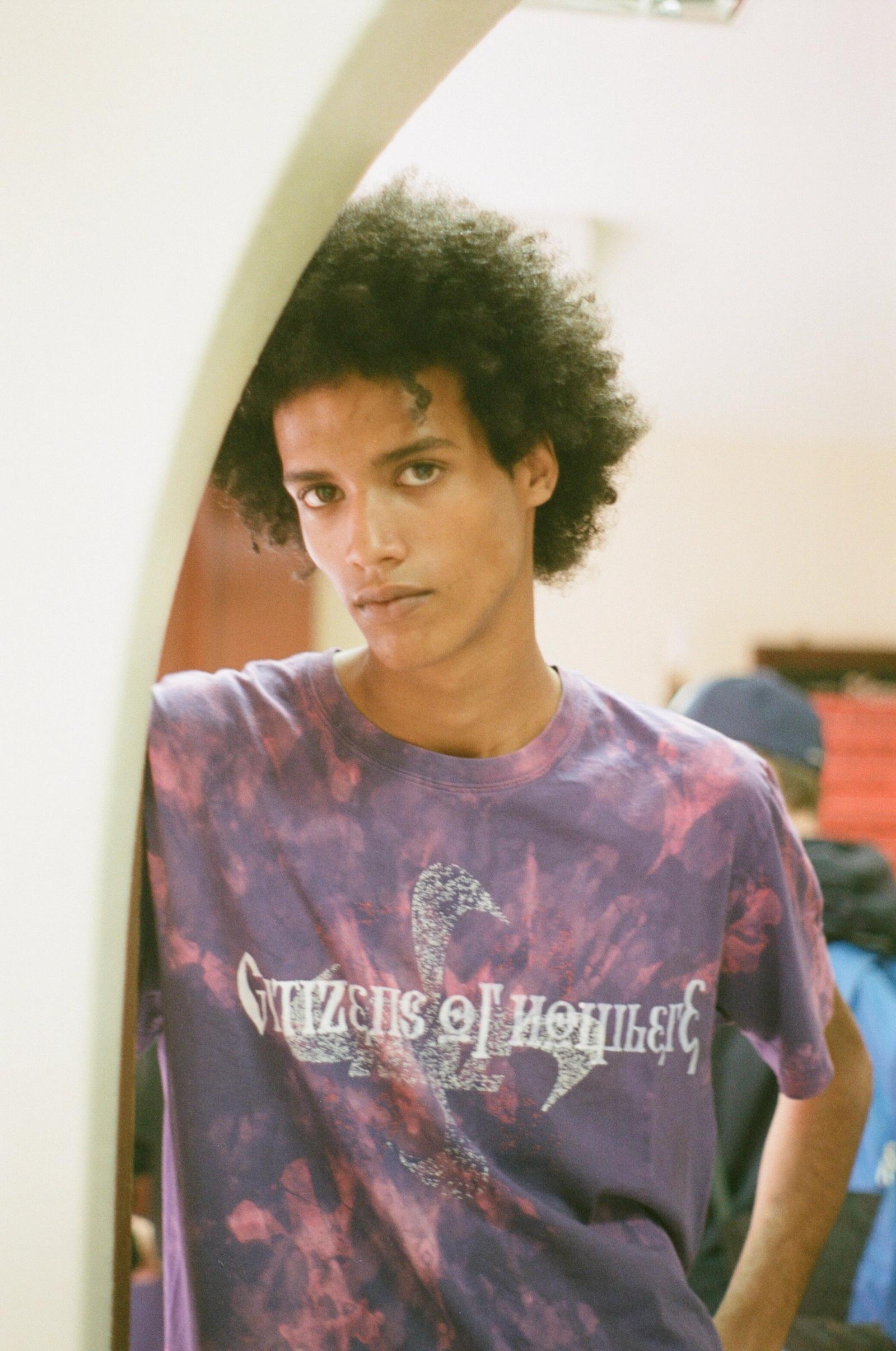 A young man with curly hair, in a purple tie-dye Citizens of Nowhere t-shirt, leans on an arched doorway and looks at the camera.