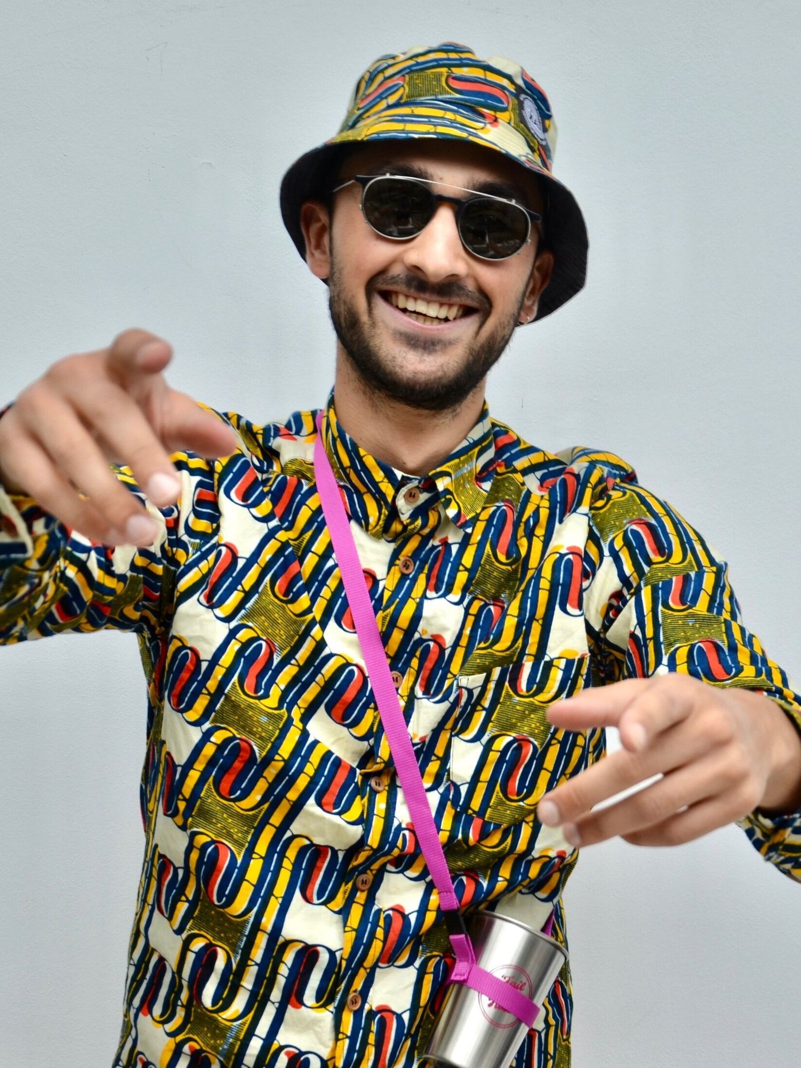 A smiling man in a bucket hat, sunglasses and a bold print shirt points at the camera, a steel cup hanging from a pink strap.