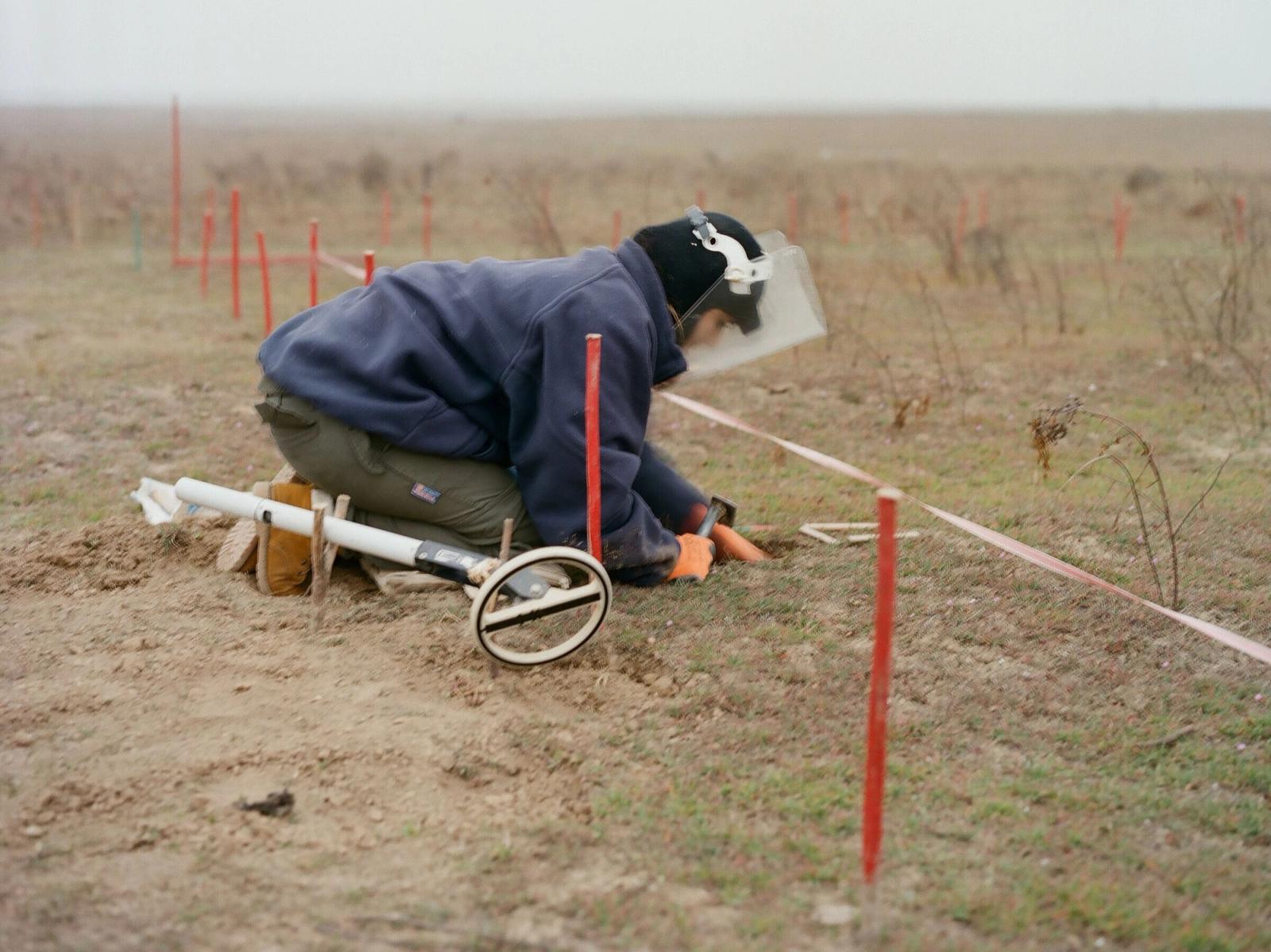 A deminer in a visor and navy fleece kneels in an open field, probing the ground beside red marker stakes and tape.