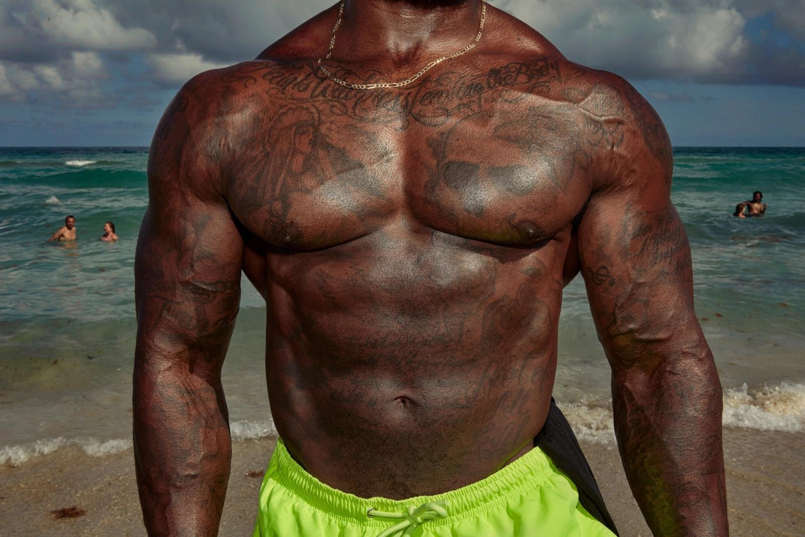 Close crop of a muscular, tattooed man's torso in neon-green swim shorts, with the sea behind him.