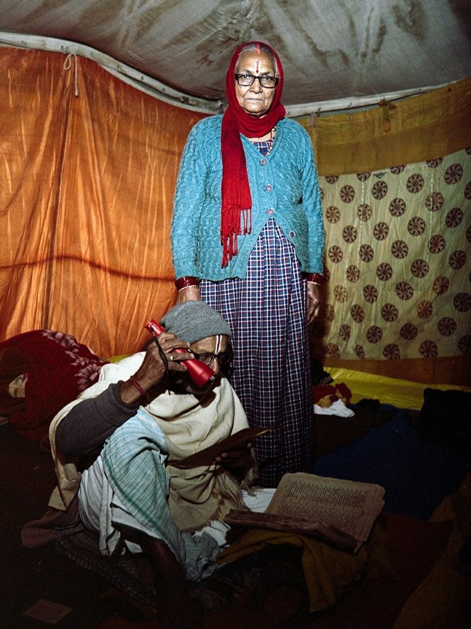 An older woman in a teal cardigan, red headscarf and glasses stands behind a seated man in a grey cap, inside an orange tent.