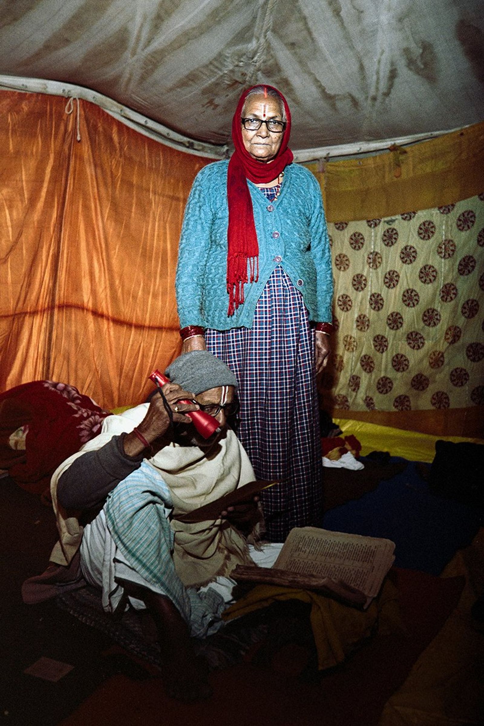 Inside a tent, an older woman in a red scarf and blue cardigan stands behind a man reading a book by torchlight.